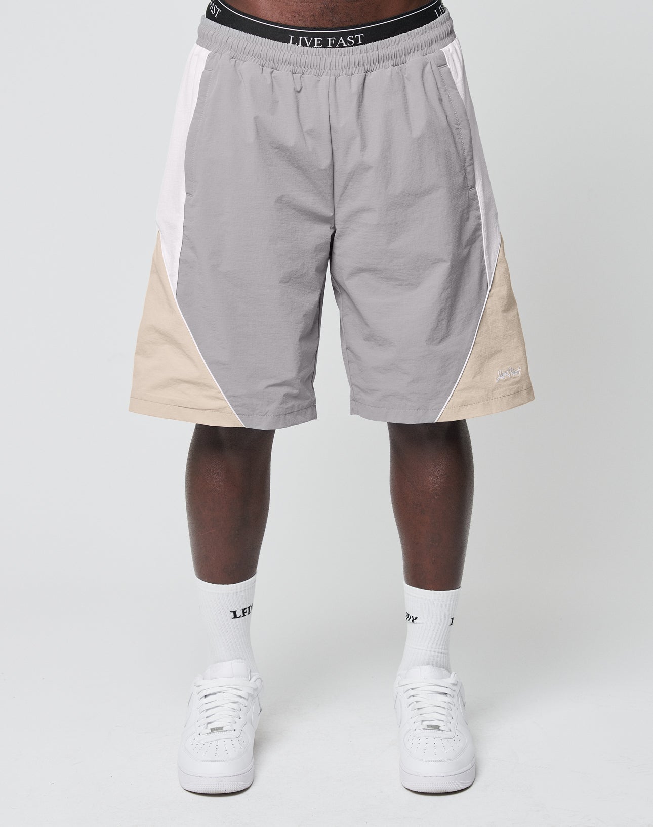 A person stands against a light gray background wearing LIVE FAST DIE YOUNG Basic Nylon Trackshorts in gray and beige with an elastic waistband, white crew socks featuring LFTD text, and white sneakers.
