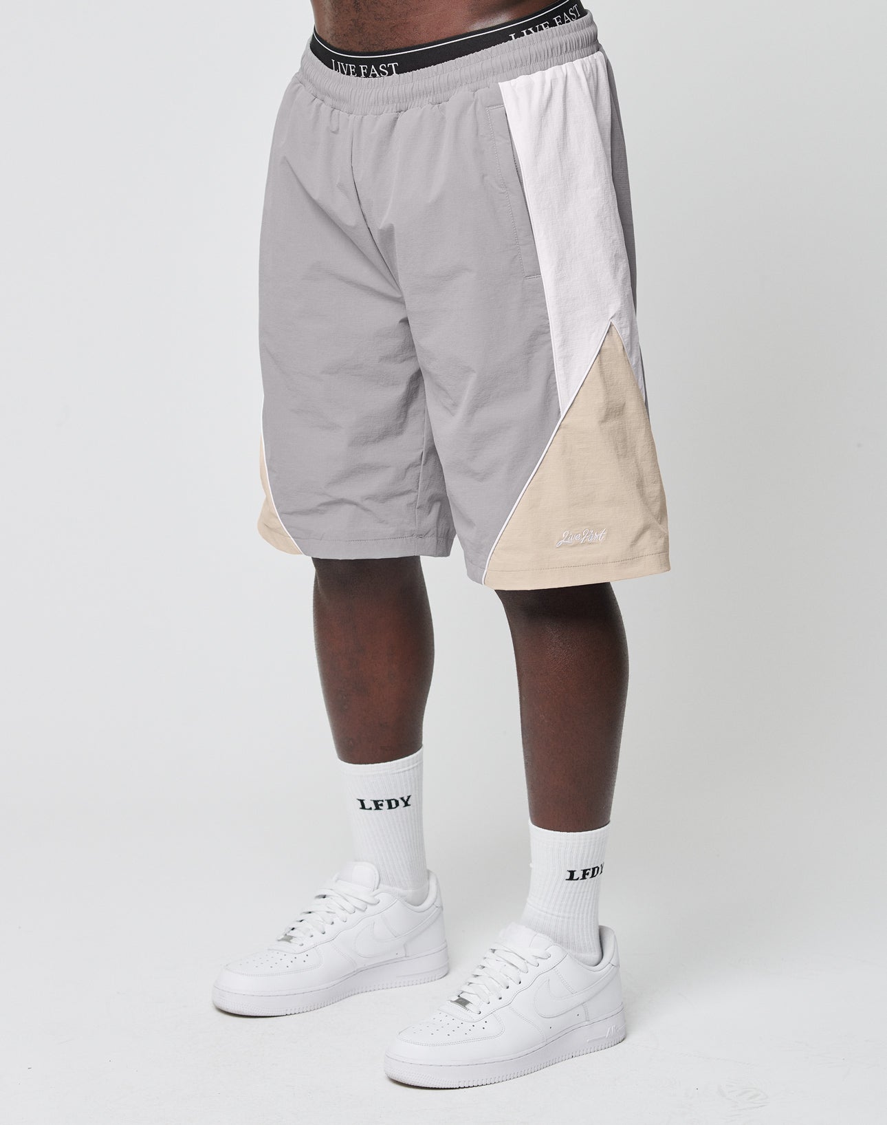 A person wearing LIVE FAST DIE YOUNG Basic Nylon Trackshorts in gray, white, and beige colorblock with a Regular Fit and elastisches Bündchen, plus white crew socks with LFDY text and white Nike Air Force 1s, stands against a plain white background.