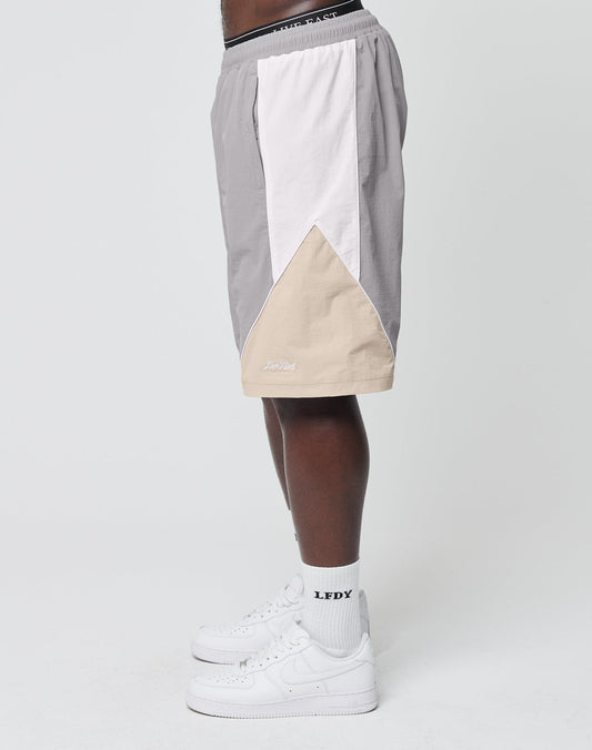 A person wears LIVE FAST DIE YOUNG Basic Nylon Trackshorts in color-block gray, white, and beige with an elastic waistband and regular fit, paired with white LFDY socks and white sneakers, standing sideways against a plain white background.