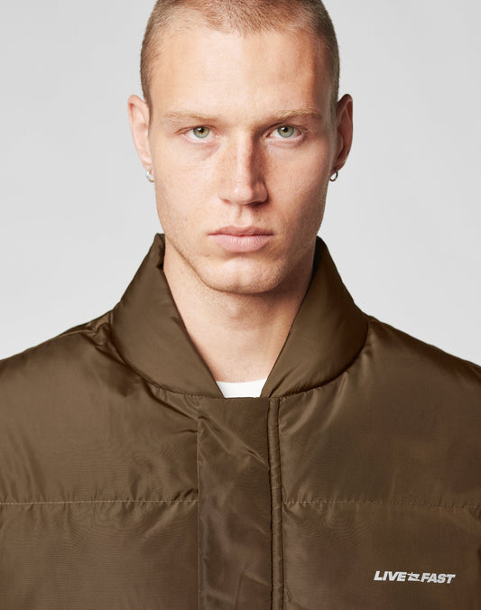 A serious-looking man with closely cropped blond hair and green eyes wears the LIVE FAST DIE YOUNG Live Fast Cropped Vest—a brown regular fit vest with white LIVE FAST text and large patch pockets. He faces the camera, wearing small hoop earrings.