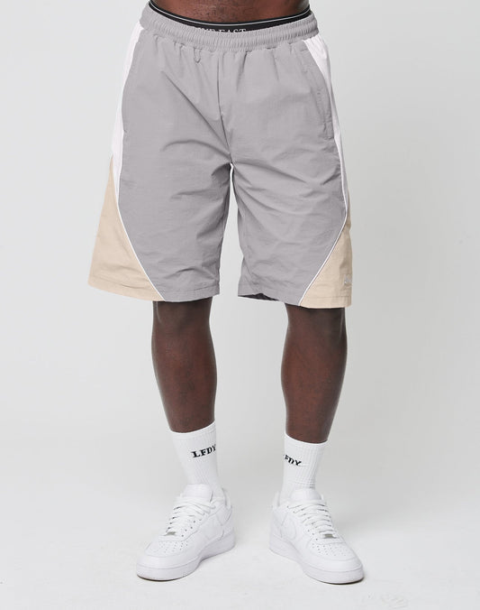A person in LIVE FAST DIE YOUNG Basic Nylon Trackshorts—gray, regular fit with beige and white side panels—paired with white LFDY logo socks and white sneakers, stands against a plain light background.