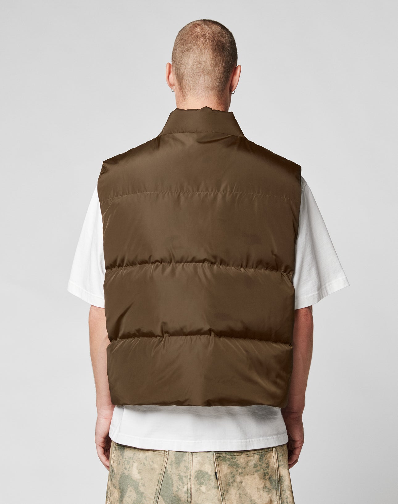A person with short hair, seen from behind, wears the LIVE FAST DIE YOUNG Live Fast Cropped Vest in brown over a white T-shirt and light-patterned pants. The background is plain light gray.