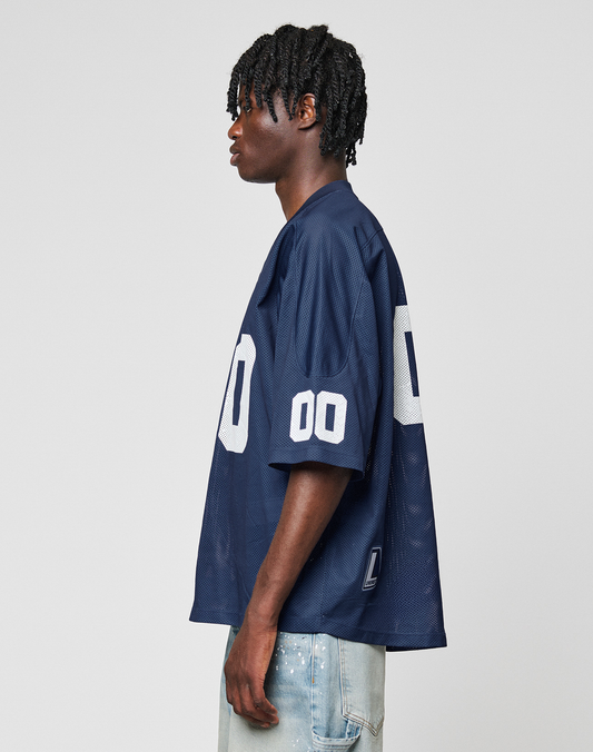 A man with short, twisted hair stands in profile against a plain white background, wearing the LIVE FAST DIE YOUNG LFDY Mesh Jersey in navy blue with the number 00 on the sleeve and back, paired with light blue jeans.