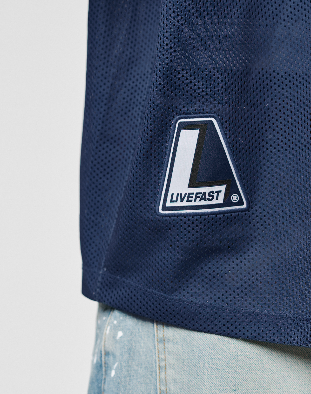 Close-up of the LFDY Mesh Jersey by LIVE FAST DIE YOUNG in dark blue, oversize fit with a V-neck and LIVEFAST patch, styled with light blue jeans featuring white paint splatters. Made from breathable polyester for comfort and style.