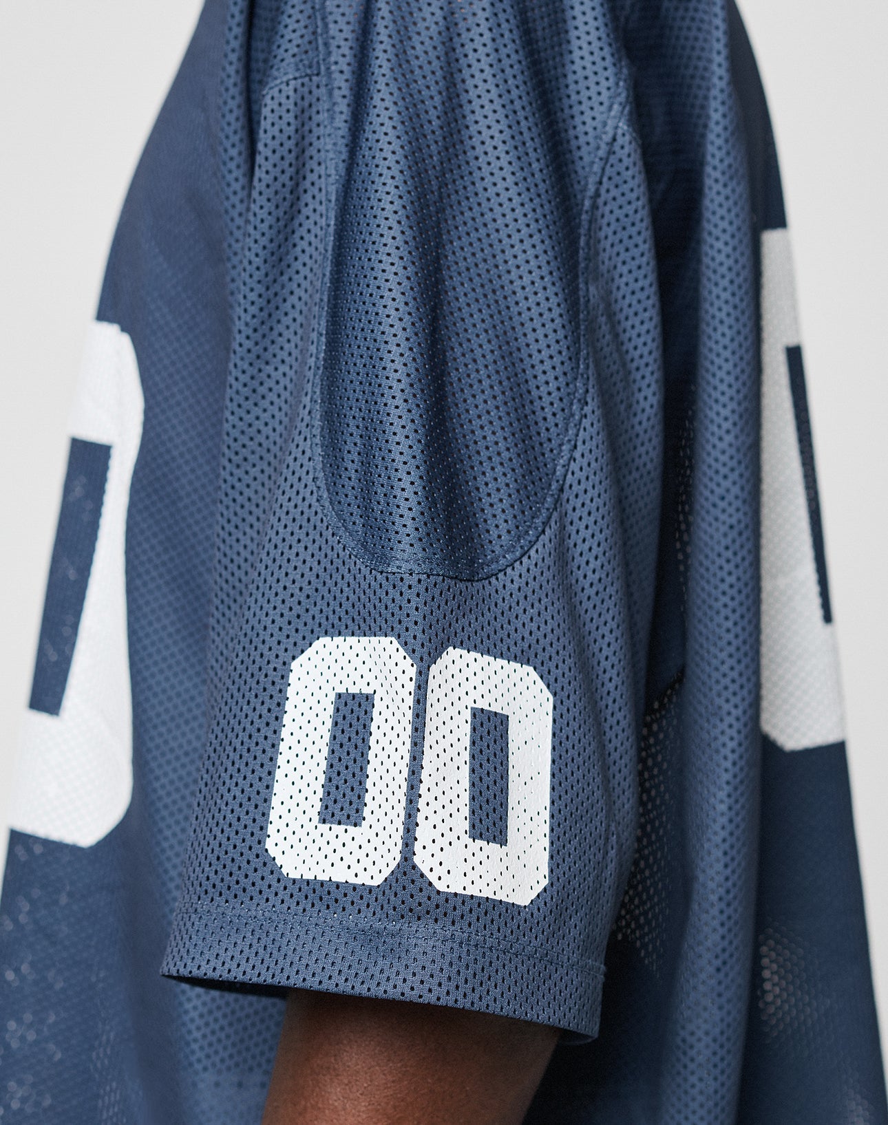 A close-up of the LFDY Mesh Jersey by LIVE FAST DIE YOUNG in navy blue polyester mesh, featuring an oversize fit and white 00 on the sleeve. The wearer’s arm is shown against a plain background.