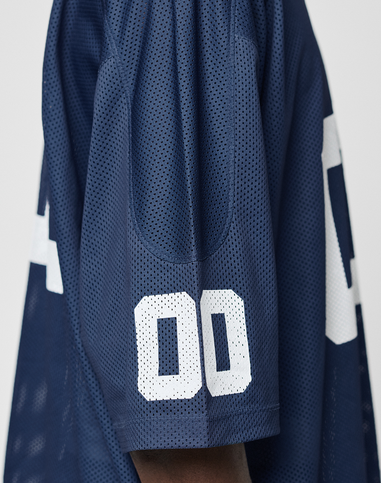 Close-up of a person wearing the LFDY Mesh Jersey by LIVE FAST DIE YOUNG, featuring an oversize fit, navy blue color with white mesh, a V-Ausschnitt, and the number 00 on the sleeve; only part of the upper arm and shoulder is shown.