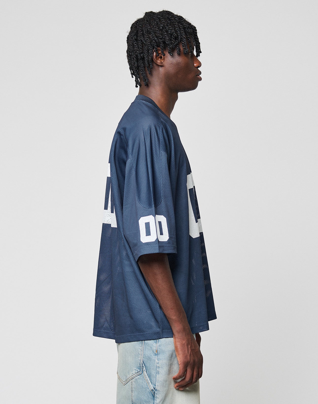 A person with short, twisted hair stands in profile wearing a loose, navy LFDY Mesh Jersey by LIVE FAST DIE YOUNG with a V-neck, white 00 numbers on the sleeve, and light blue jeans. The background is plain white.