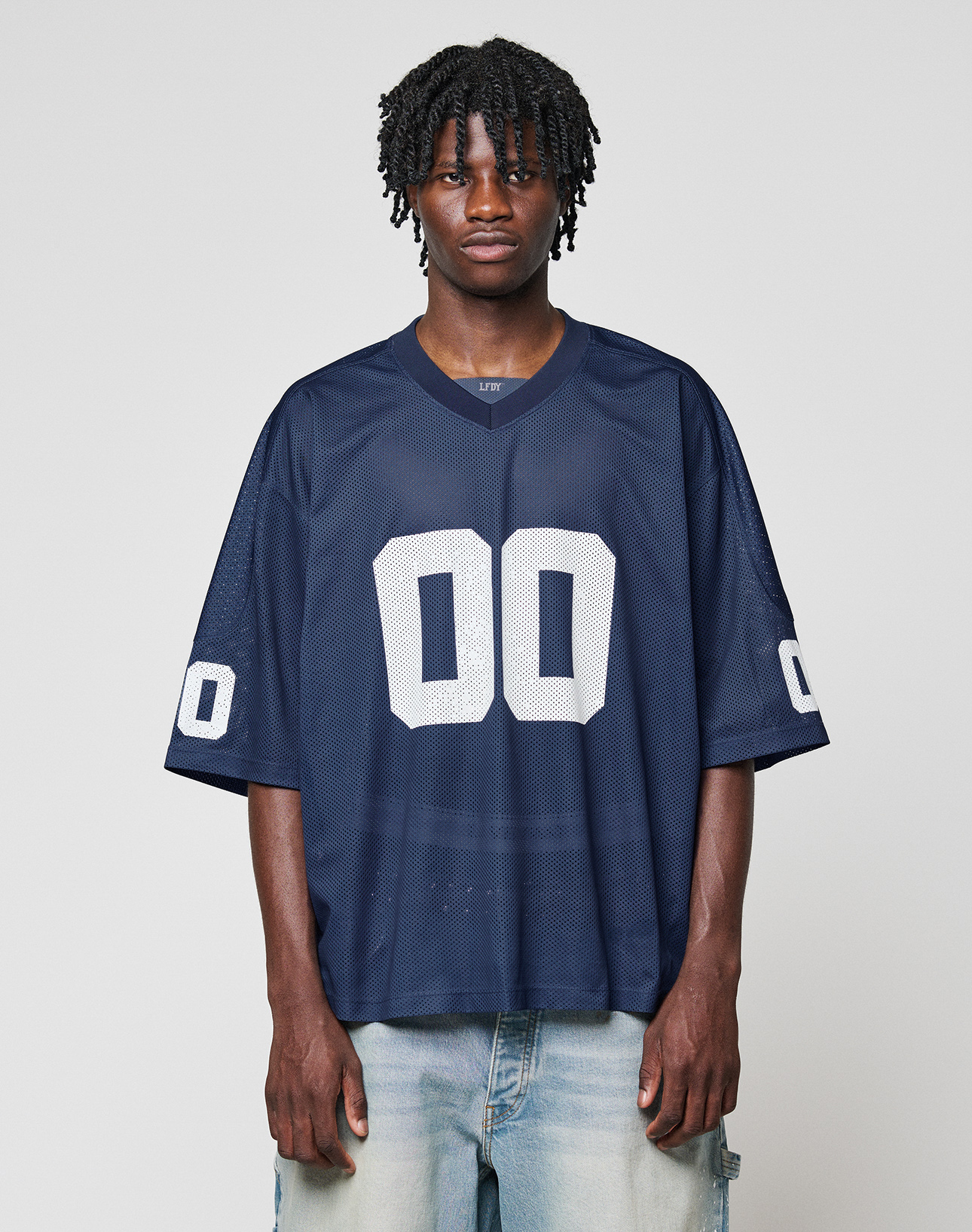 A person wears a loose-fitting LIVE FAST DIE YOUNG LFDY Mesh Jersey in navy blue with a V-neck and white number 00 on the front and sleeves, paired with light blue jeans, standing against a plain light gray background. | model