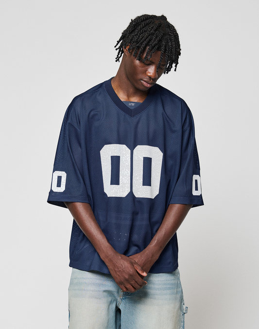 A person wearing the oversized navy blue LFDY Mesh Jersey by LIVE FAST DIE YOUNG, featuring a V-Ausschnitt and number 00 on the front and sleeves, stands with hands clasped, looking down against a plain light gray background.
