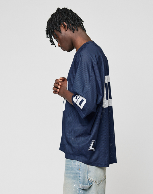 A person with short, twisted hair stands in profile wearing the LIVE FAST DIE YOUNG LFDY Mesh Jersey in navy blue with V-Ausschnitt and Oversize Fit, paired with light blue jeans, hands clasped against a plain light background.