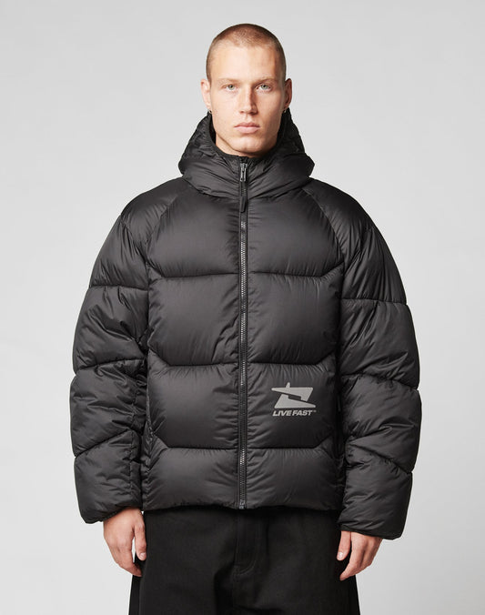 A person with short hair wears the LIVE FAST DIE YOUNG Live Fast Puffer Jacket—black, hooded, water-resistant, and featuring a reflective LIVE FAST print on the left chest—against a plain light gray background.