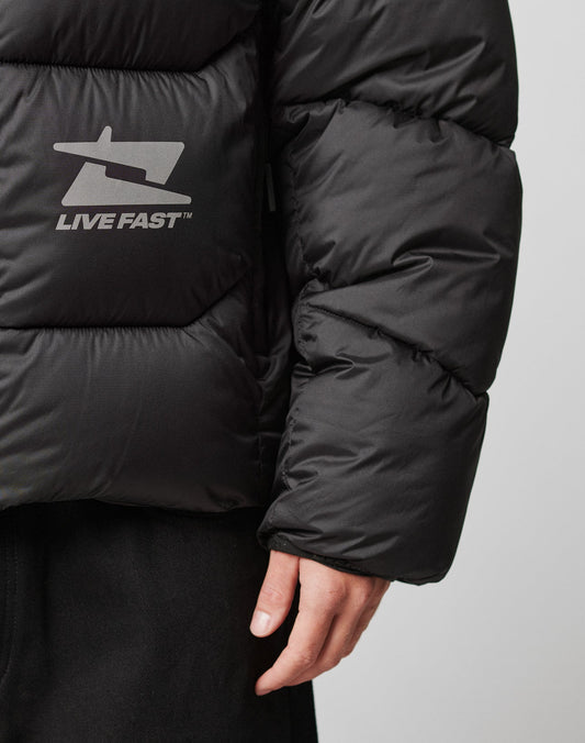 A person wearing the LIVE FAST DIE YOUNG Live Fast Puffer Jacket in black, featuring an oversize fit with LIVE FAST and a logo in reflective print, with only their arm and hand visible against a plain background.