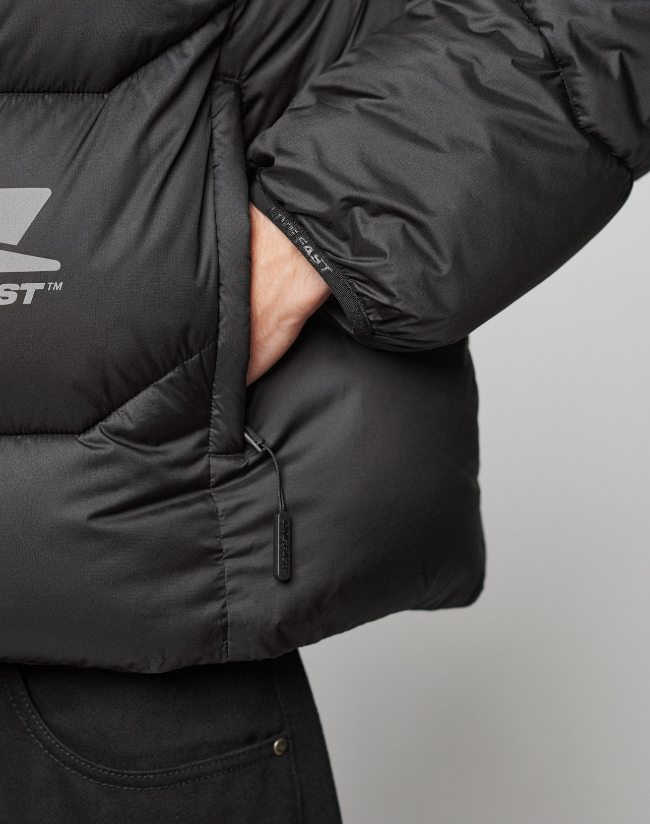 A close-up shows a person wearing the LIVE FAST DIE YOUNG Live Fast Puffer Jacket in black, with their hand in the pocket; the water-repellent jacket features a visible zipper pull and part of black pants.