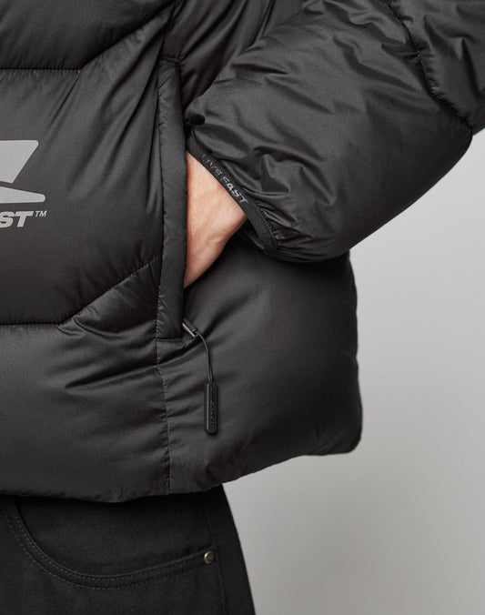 A close-up shows a person wearing the LIVE FAST DIE YOUNG Live Fast Puffer Jacket in black, with their hand in the pocket; the water-repellent jacket features a visible zipper pull and part of black pants.