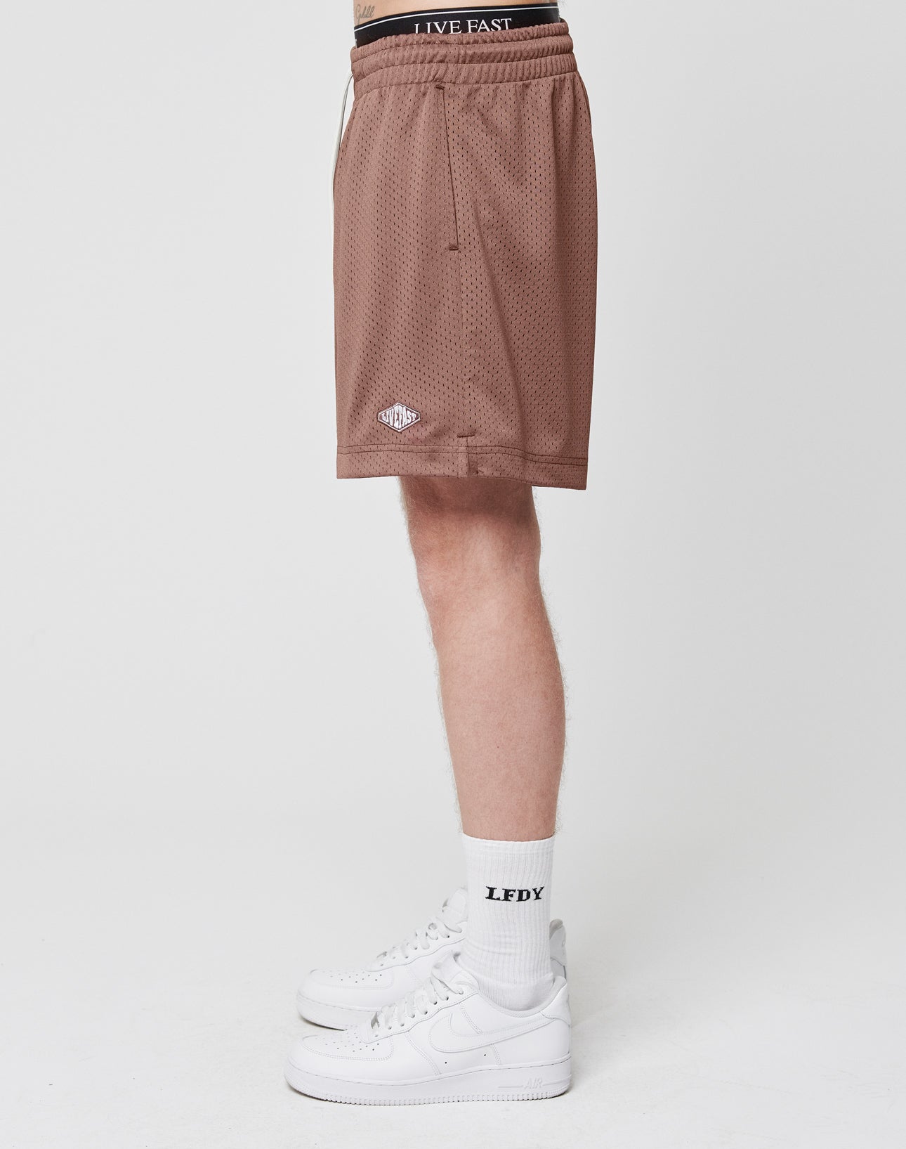 A person stands in profile against a plain white background wearing LIVE FAST DIE YOUNG Basic Mesh Shorts in brown, LFDY-branded white socks, and white sneakers.