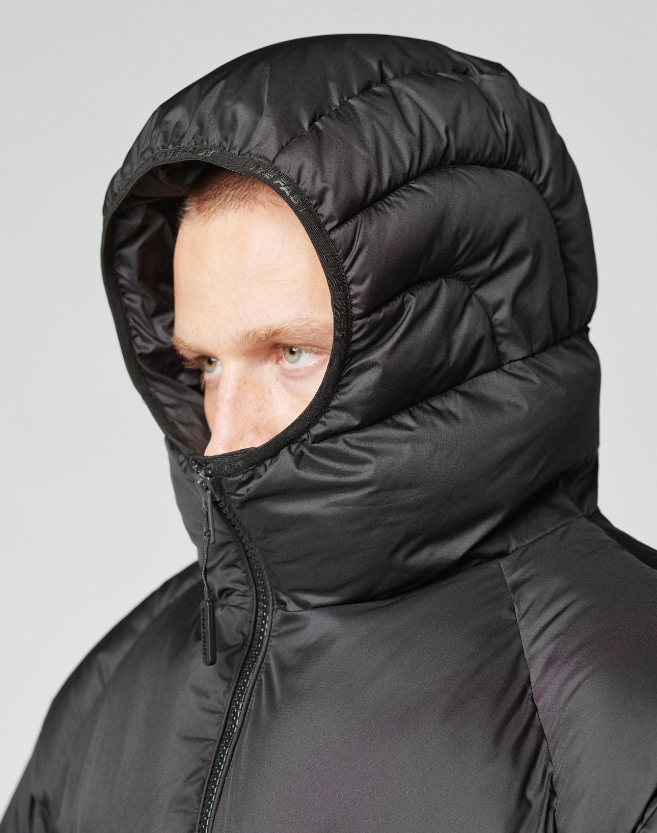 A person wears the LIVE FAST DIE YOUNG Live Fast Puffer Jacket in black, hood up and zipped to the chin, with an oversized fit partially covering their face as they look to the side against a plain background.