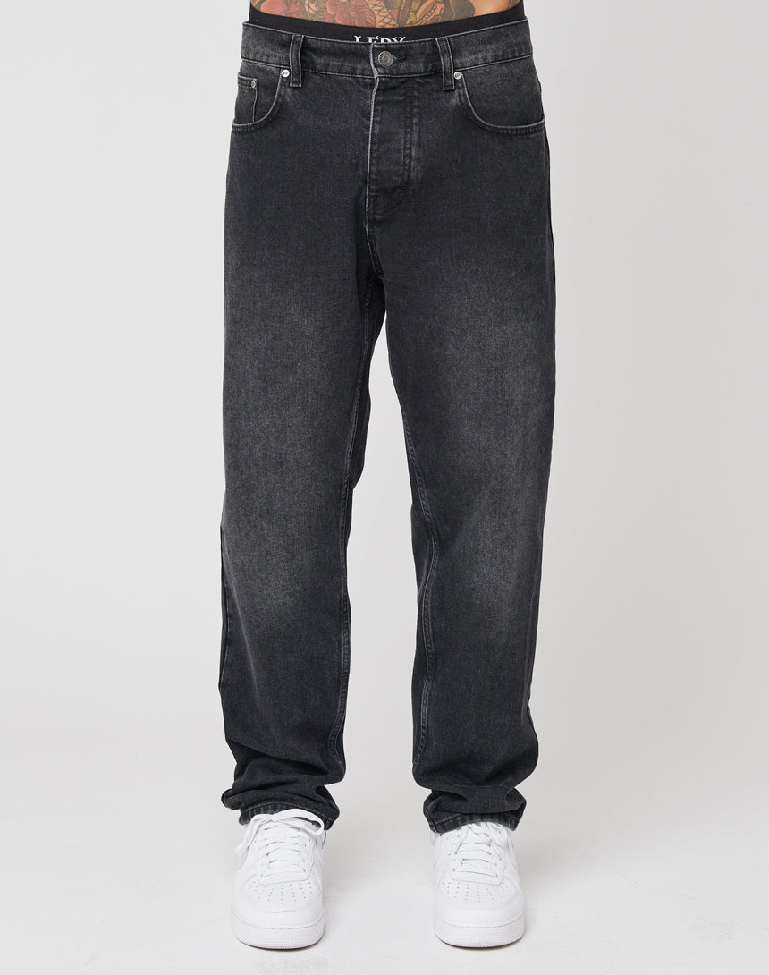 A person wearing LIVE FAST DIE YOUNG Regular Denim black washed jeans with a distinctive Lederpatch and white sneakers stands against a plain white background, only their lower half visible.