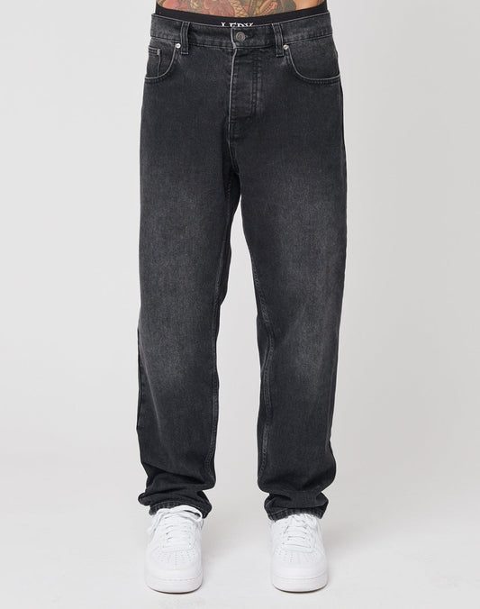A person wearing LIVE FAST DIE YOUNG Regular Denim black washed jeans with a distinctive Lederpatch and white sneakers stands against a plain white background, only their lower half visible.