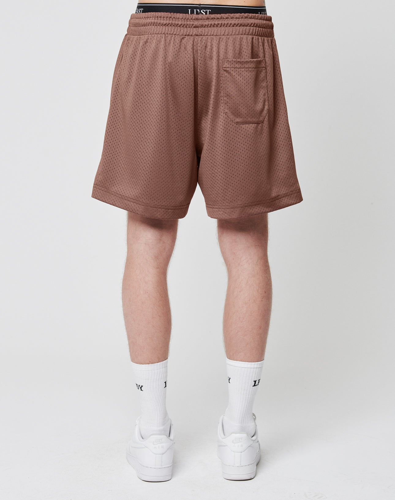 A person stands facing away, wearing LIVE FAST DIE YOUNG Basic Mesh Shorts in brown with a logo patch, elastic waistband, and back pocket, paired with white socks and sneakers. The background is plain and light-colored.