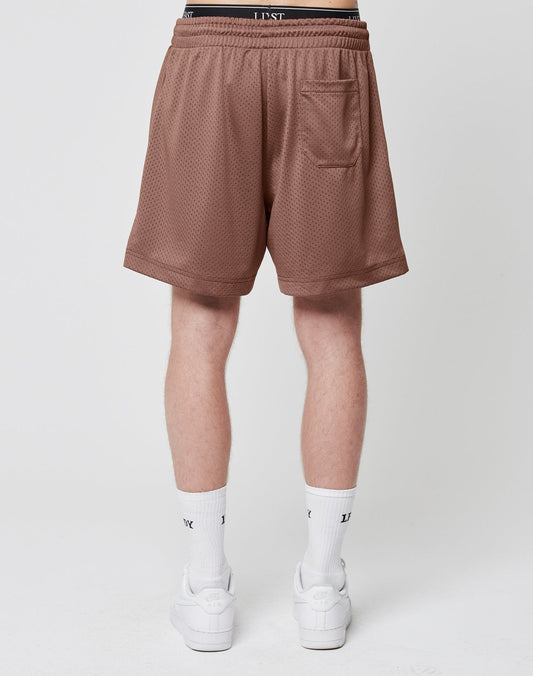 A person stands facing away, wearing LIVE FAST DIE YOUNG Basic Mesh Shorts in brown with a logo patch, elastic waistband, and back pocket, paired with white socks and sneakers. The background is plain and light-colored.
