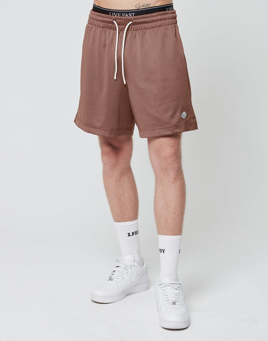 A person stands against a plain white background wearing LIVE FAST DIE YOUNG Basic Mesh Shorts in light brown, white socks with black text, white sneakers, and a black waistband with white lettering and a logo patch. The upper body is not visible.