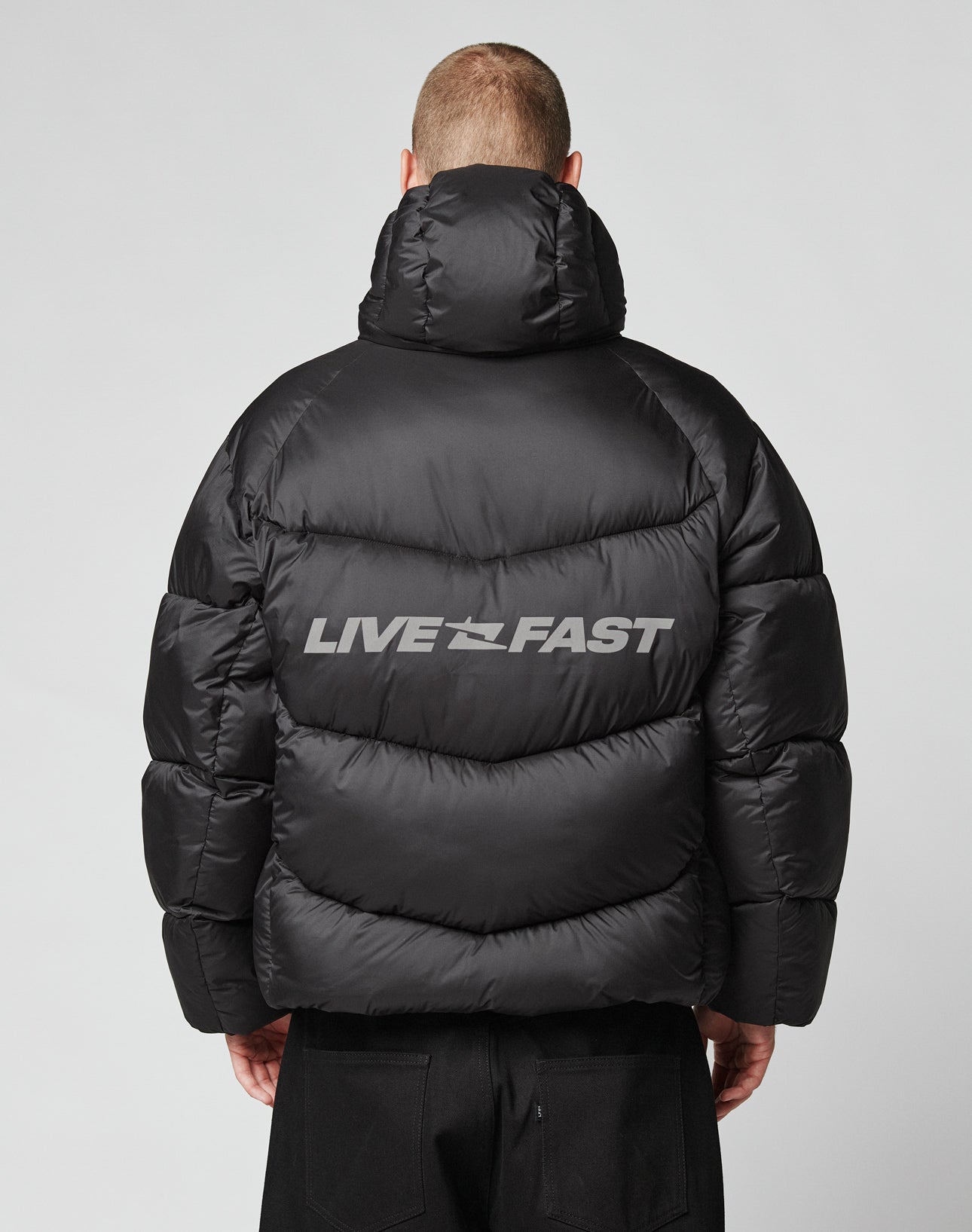 Someone faces away from the camera in a shiny black Live Fast Puffer Jacket by LIVE FAST DIE YOUNG, featuring an oversized fit and bold gray reflective LIVE FAST print across the back, standing against a plain light background.