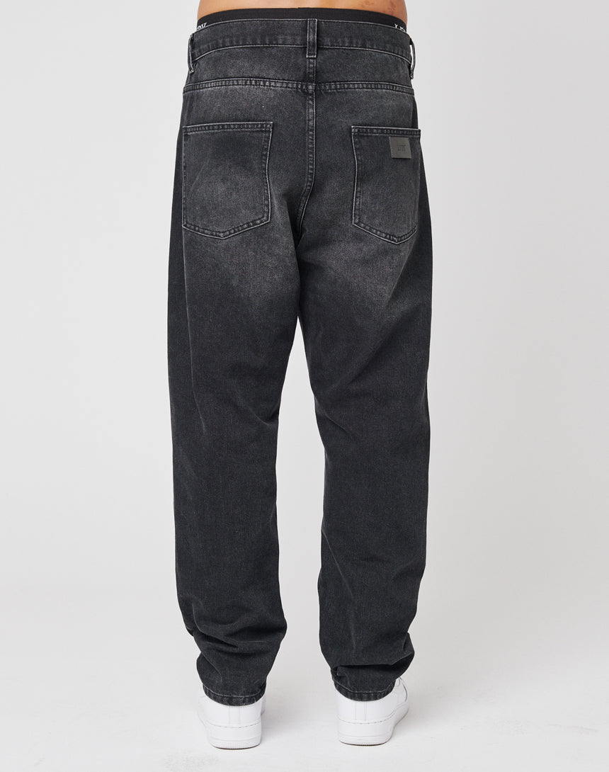 A person is shown from the back wearing LIVE FAST DIE YOUNG Regular Denim jeans—loose-fitting, faded black, 100% Baumwolle—with two large back pockets, visible stitching, a subtle Lederpatch, and white sneakers against a plain background.