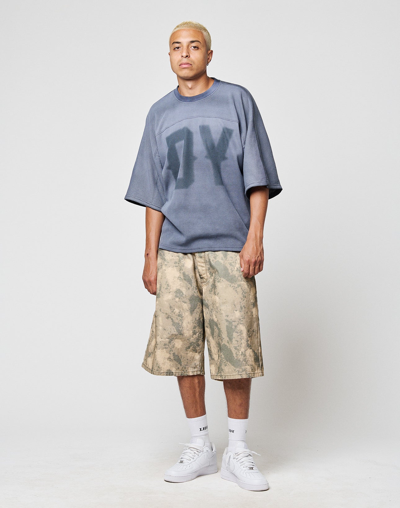 A person with short blond hair wears a blue Faded Waffle Jersey from LIVE FAST DIE YOUNG, beige camo knee-length shorts, white socks, and white sneakers, standing against a plain light background.