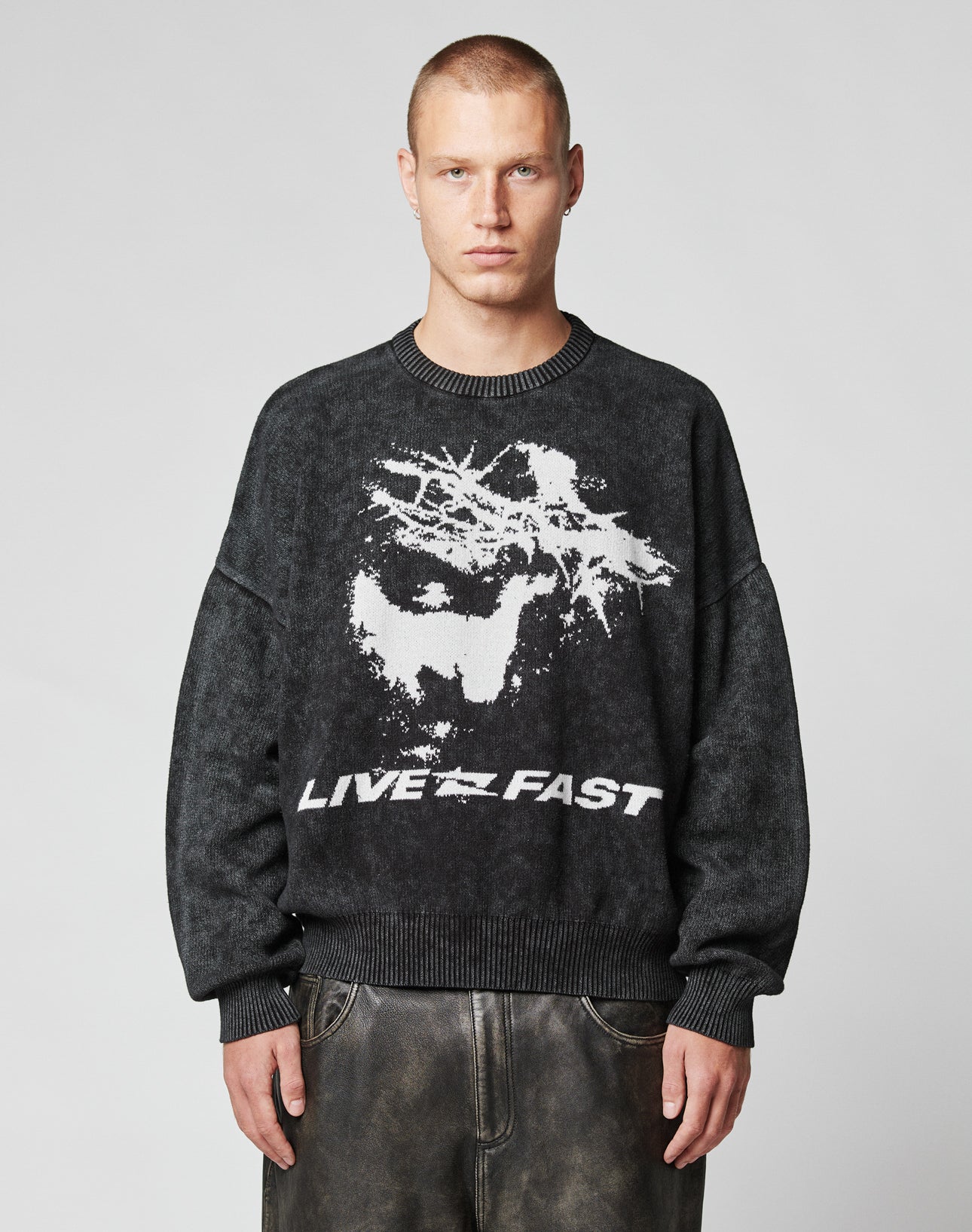 A person with short hair wears the LFDY Savior Knit Sweater by LIVE FAST DIE YOUNG—an oversized dark sweater with a bold abstract white graphic and LIVE FAST text, standing against a plain light background.