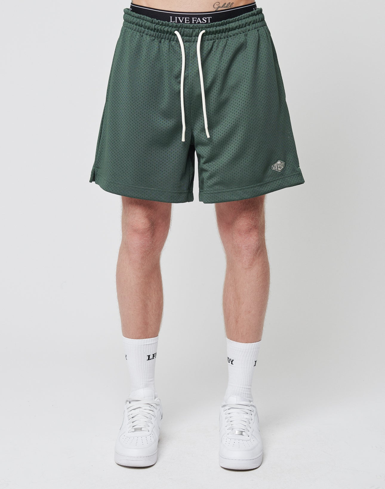 A person wears LIVE FAST DIE YOUNG Basic Mesh Shorts in green with a white drawstring and logo patch, paired with white socks and sneakers, standing against a plain white background.