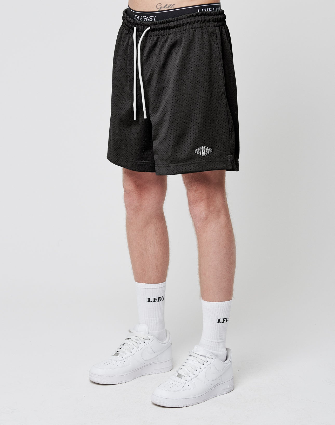 Wearing LIVE FAST DIE YOUNG Basic Mesh Shorts in black with a white drawstring and logo patch, paired with white LFDY crew socks and Nike sneakers, standing against a plain white background. Upper body is not visible.