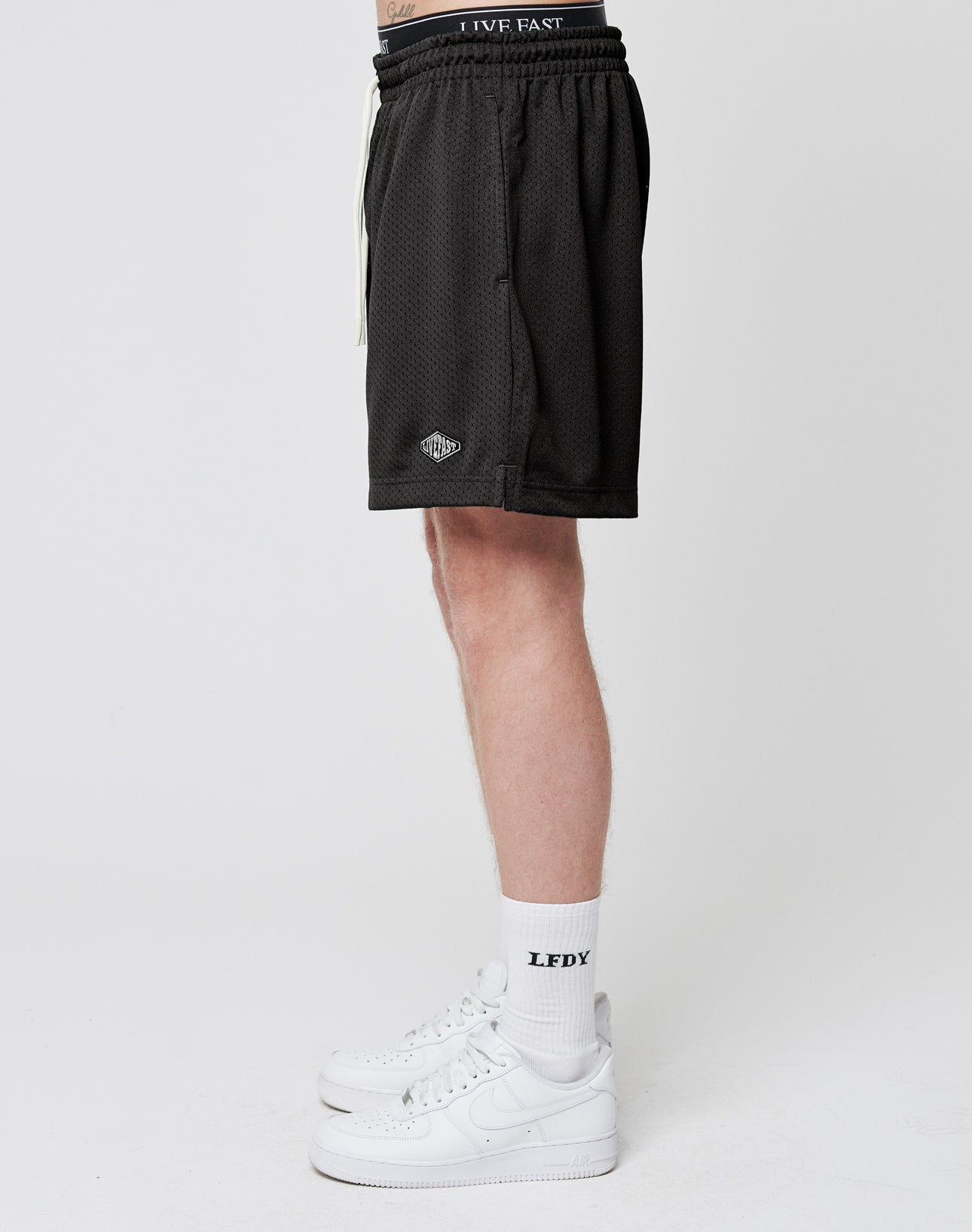 A person stands in profile against a white background wearing LIVE FAST DIE YOUNG Basic Mesh Shorts, white LFDY socks, and white sneakers. Only their legs and part of their torso are visible.