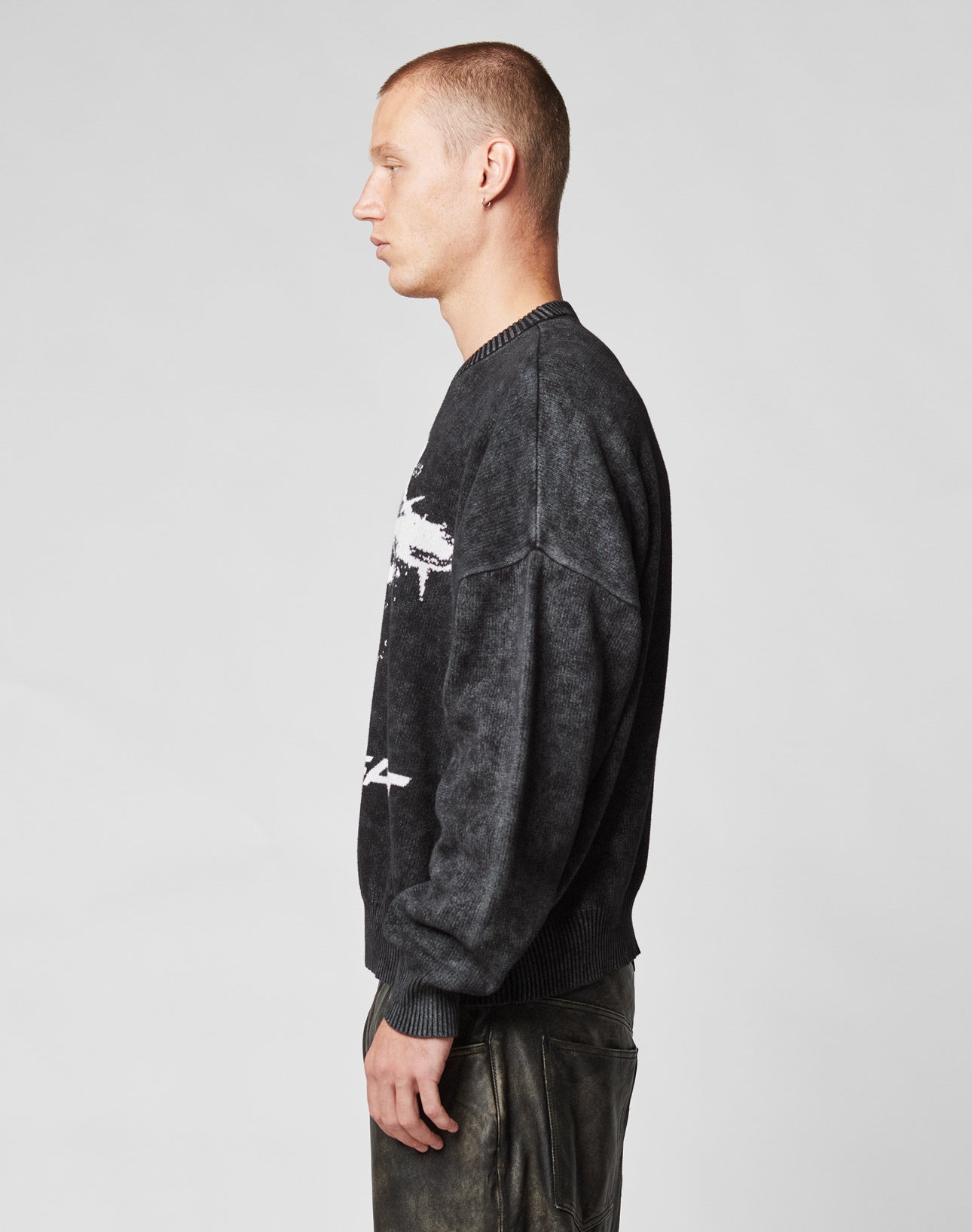 A young man with short hair stands in profile against a plain background, wearing the LFDY Savior Knit Sweater by LIVE FAST DIE YOUNG and dark pants.
