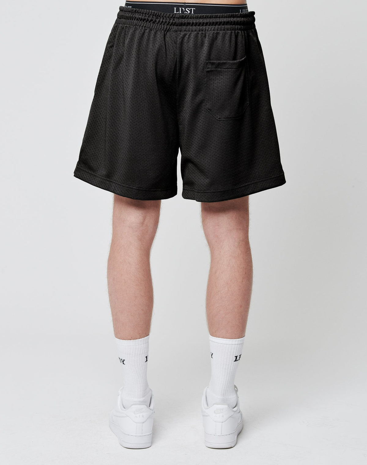 A person stands facing away, wearing LIVE FAST DIE YOUNG Basic Mesh Shorts—black polyester with an elastic waistband and one back pocket. The look is finished with white socks featuring black text and white sneakers on a plain white background.