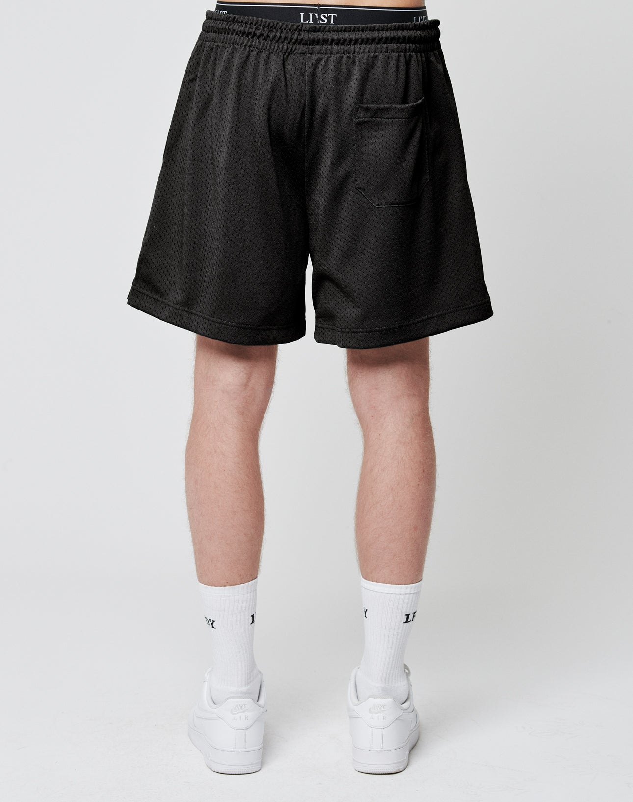 A person stands facing away, wearing LIVE FAST DIE YOUNG Basic Mesh Shorts—black polyester with an elastic waistband and one back pocket. The look is finished with white socks featuring black text and white sneakers on a plain white background.