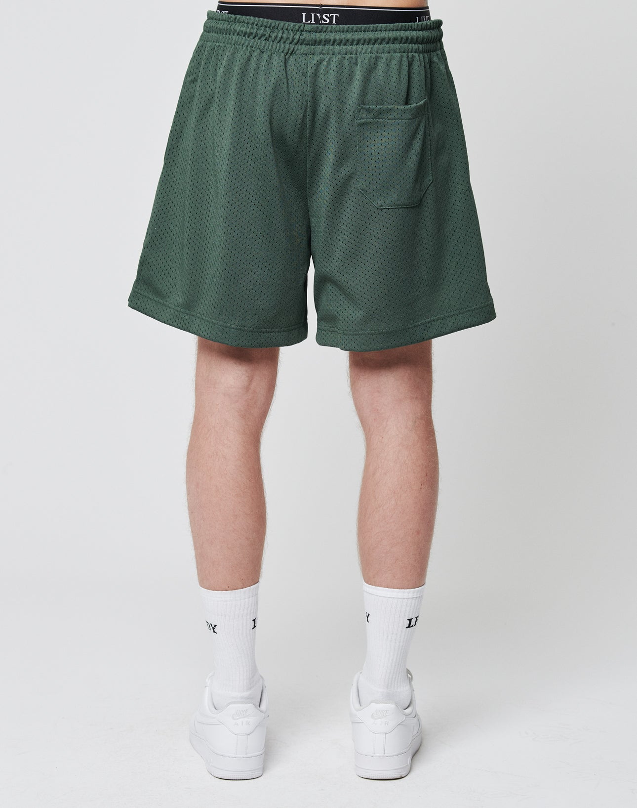 A person is shown from the waist down wearing LIVE FAST DIE YOUNG Basic Mesh Shorts in green with an elastic waistband, LJST branding, a back pocket, white socks, and white sneakers. The background is plain white.