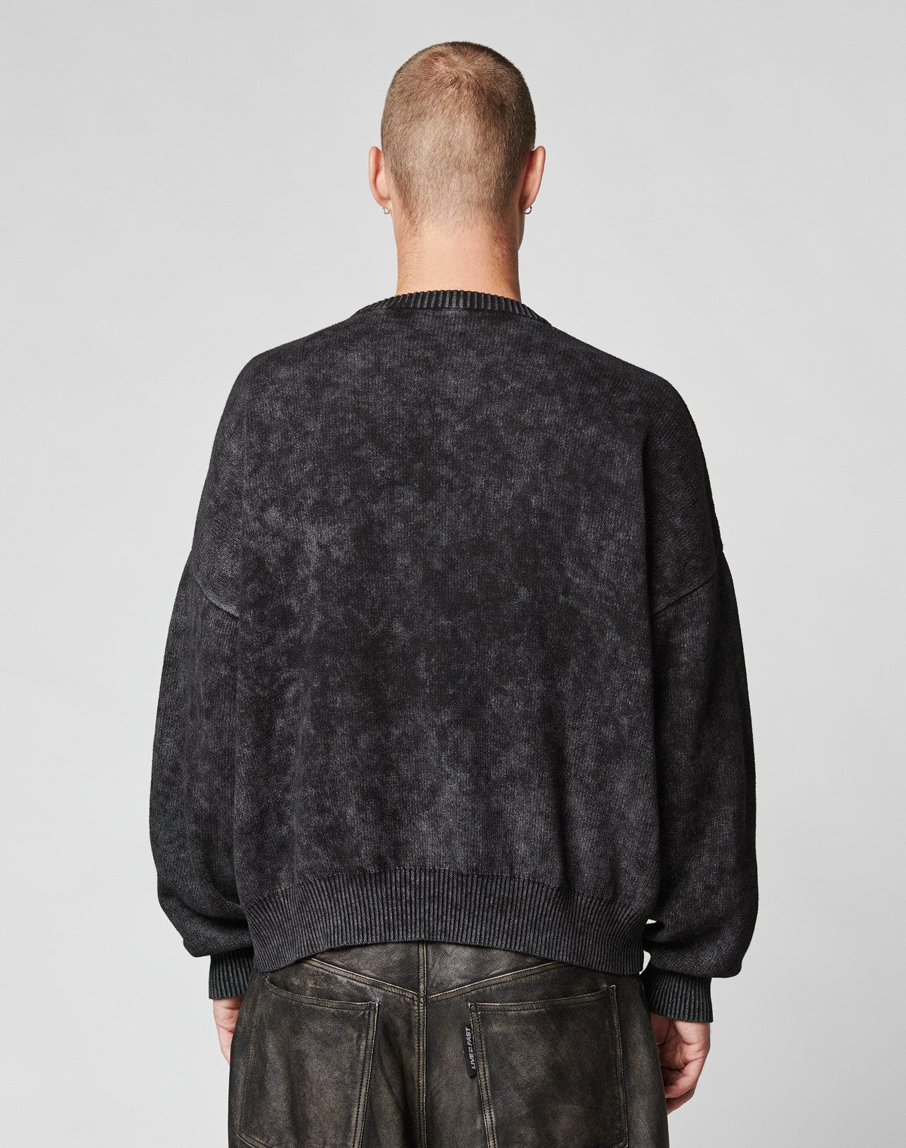 A person with a shaved head, seen from behind, wears a loose-fitting LFDY Savior Knit Sweater by LIVE FAST DIE YOUNG in dark grey and dark pants with visible seams and zippers. The background is plain and light-colored.