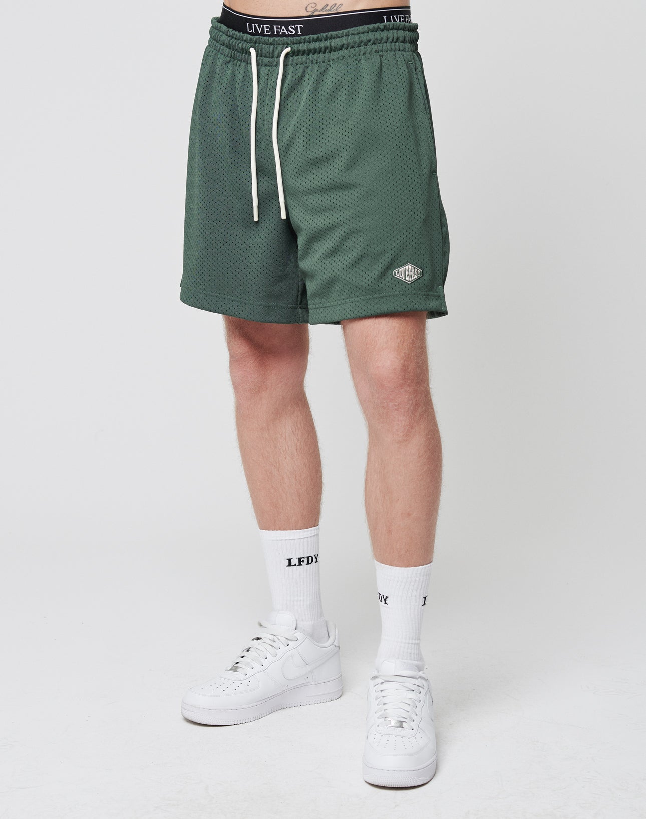 Someone is seen wearing LIVE FAST DIE YOUNG Basic Mesh Shorts in green with an elastic waistband and small logo patch, paired with LFDY-branded white crew socks and white sneakers. Only the lower half is shown; the background is plain white.