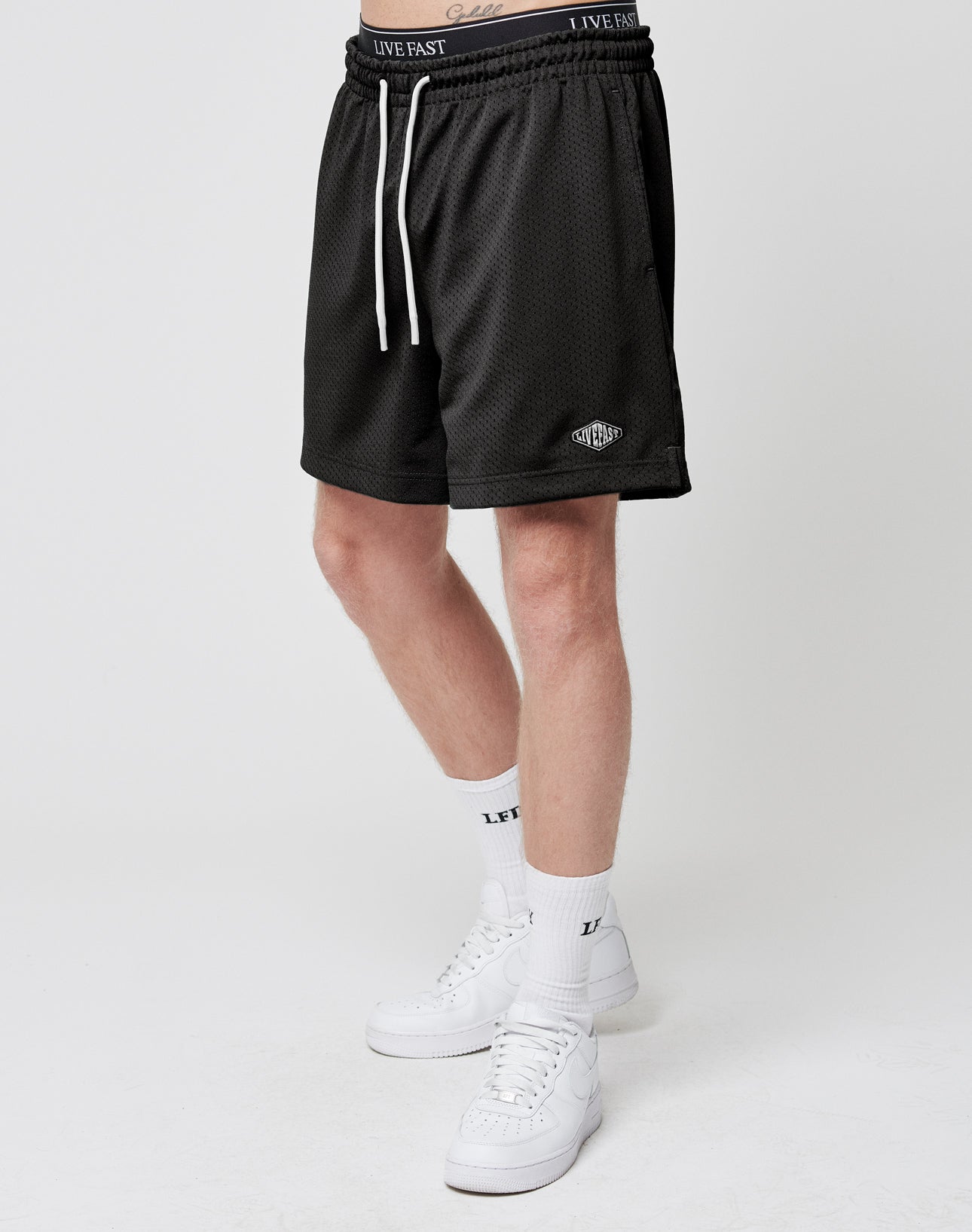 A person wears LIVE FAST DIE YOUNG Basic Mesh Shorts with an elastic waistband and logo patch near the hem, white LIVE FAST crew socks, and white sneakers, standing against a plain white background.