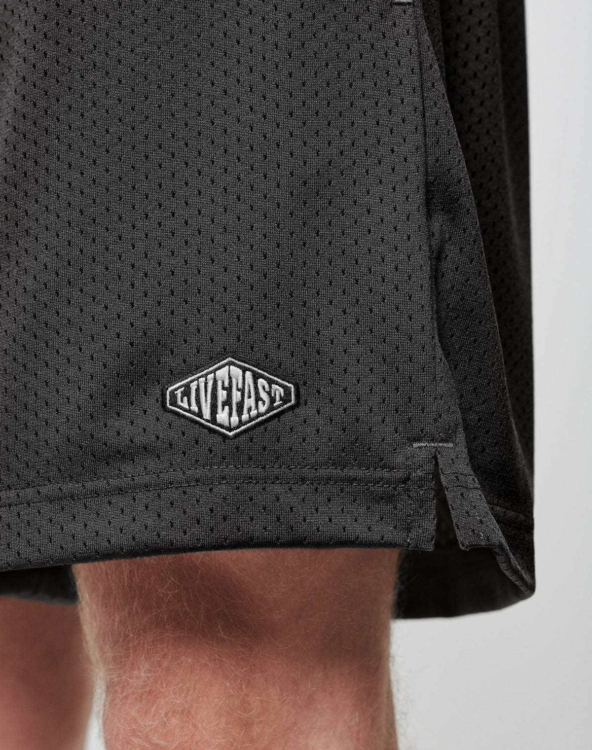 Close-up of the lower part of LIVE FAST DIE YOUNG Basic Mesh Shorts in black, featuring an elastic waistband and a white LIVE FAST logo patch on the hem, made from 100% polyester and worn by a light-skinned person.