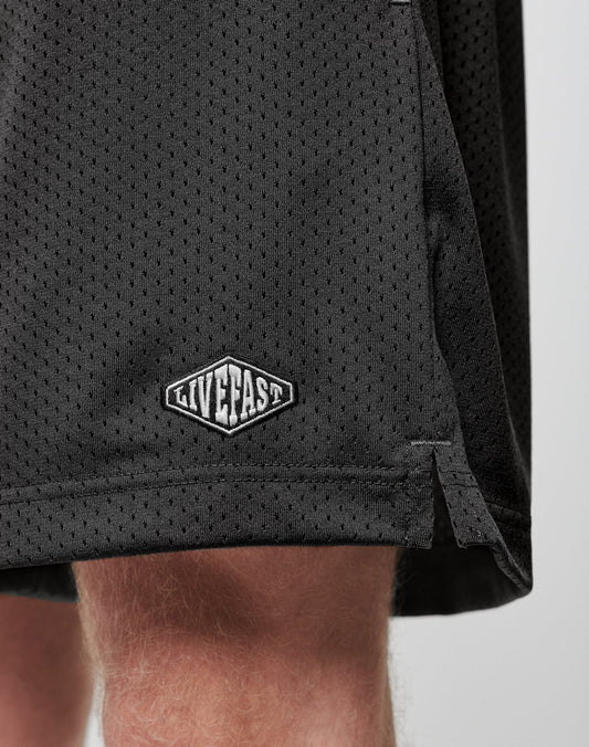 Close-up of the lower part of LIVE FAST DIE YOUNG Basic Mesh Shorts in black, featuring an elastic waistband and a white LIVE FAST logo patch on the hem, made from 100% polyester and worn by a light-skinned person.