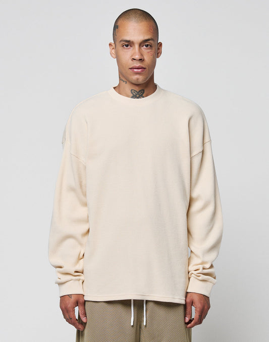 A person with a shaved head and visible neck tattoo wears the LIVE FAST DIE YOUNG Blank Waffle Longsleeve, an oversized cream 100% Baumwolle shirt, with tan shorts, standing against a plain light gray background.