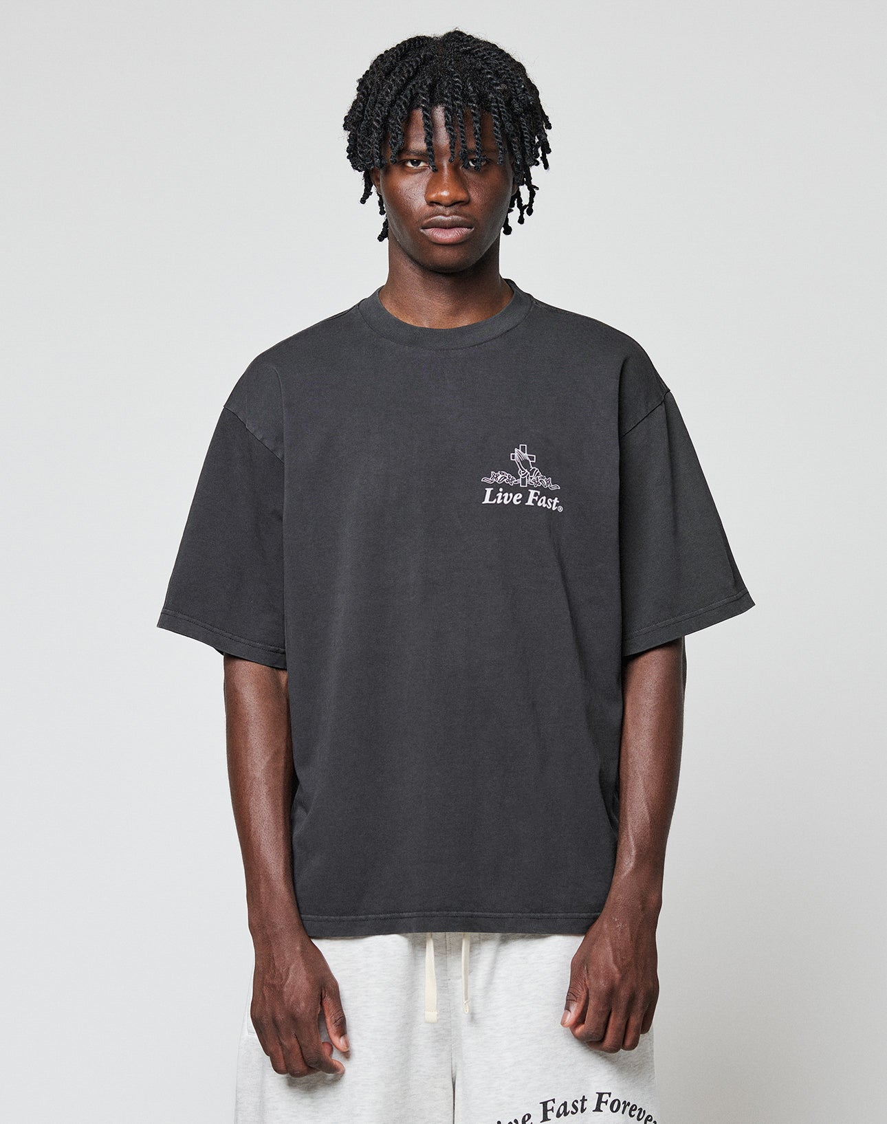 A person with short, twisted braids wears the LIVE FAST DIE YOUNG LFDY Praying Tee, a loose dark shirt featuring Live Fast text, paired with light shorts displaying Live Fast Forever on the waistband, against a plain background.