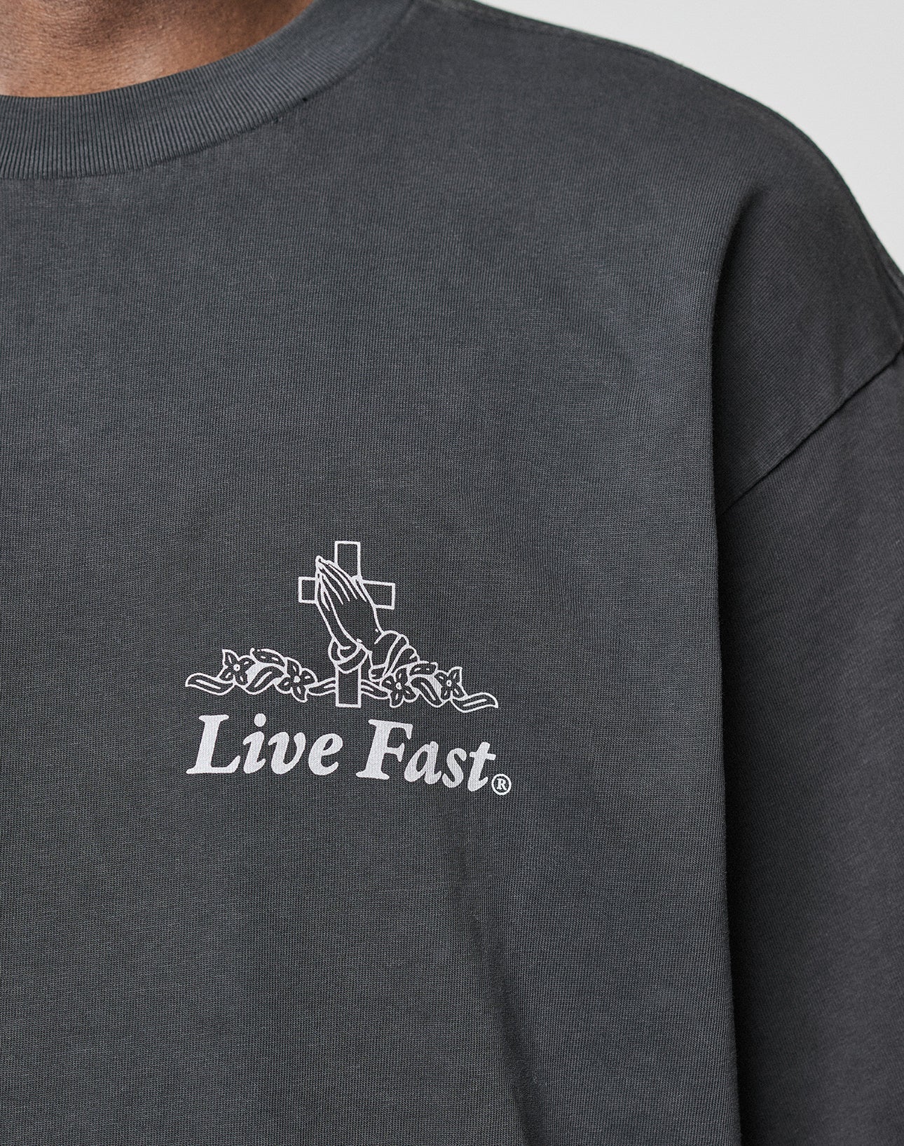 A close-up of the LFDY Praying Tee by LIVE FAST DIE YOUNG, an oversize fit dark gray shirt made from 100% cotton, with a white praying hands and cross graphic, roses, and Live Fast text below.