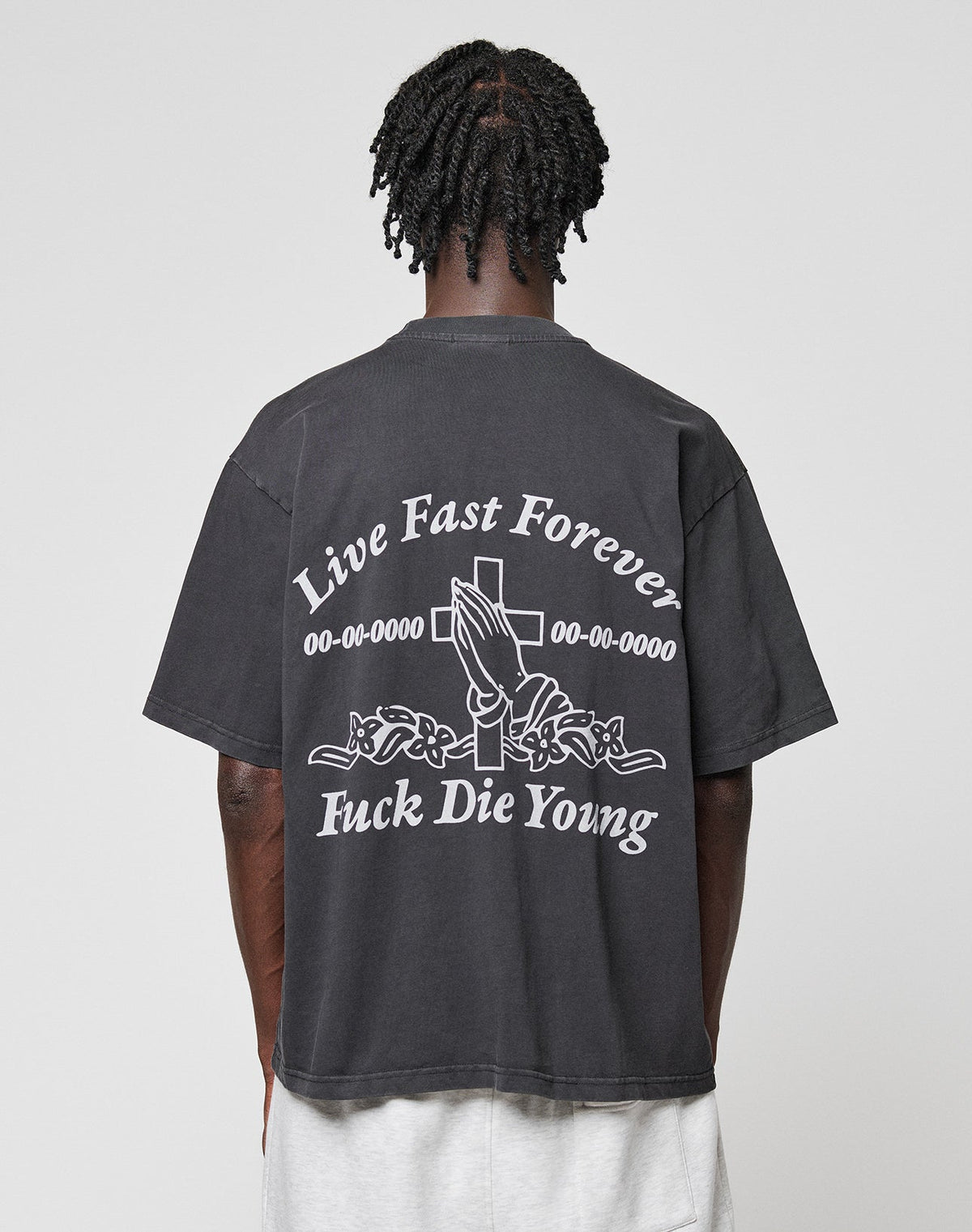 Seen from behind, a person with short, twisted hair wears the black LFDY Praying Tee by LIVE FAST DIE YOUNG, featuring white graphics of praying hands, a cross, roses, and the phrases Live Fast Forever and Fuck Die Young.| model