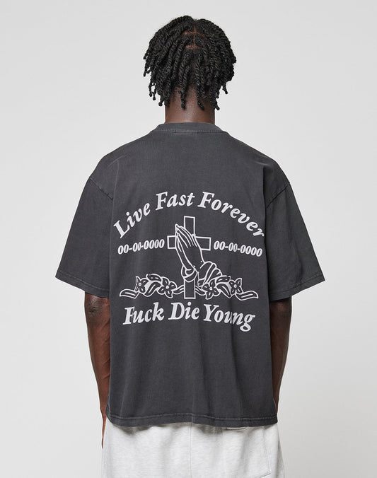 Seen from behind, a person with short, twisted hair wears the black LFDY Praying Tee by LIVE FAST DIE YOUNG, featuring white graphics of praying hands, a cross, roses, and the phrases Live Fast Forever and Fuck Die Young.| model