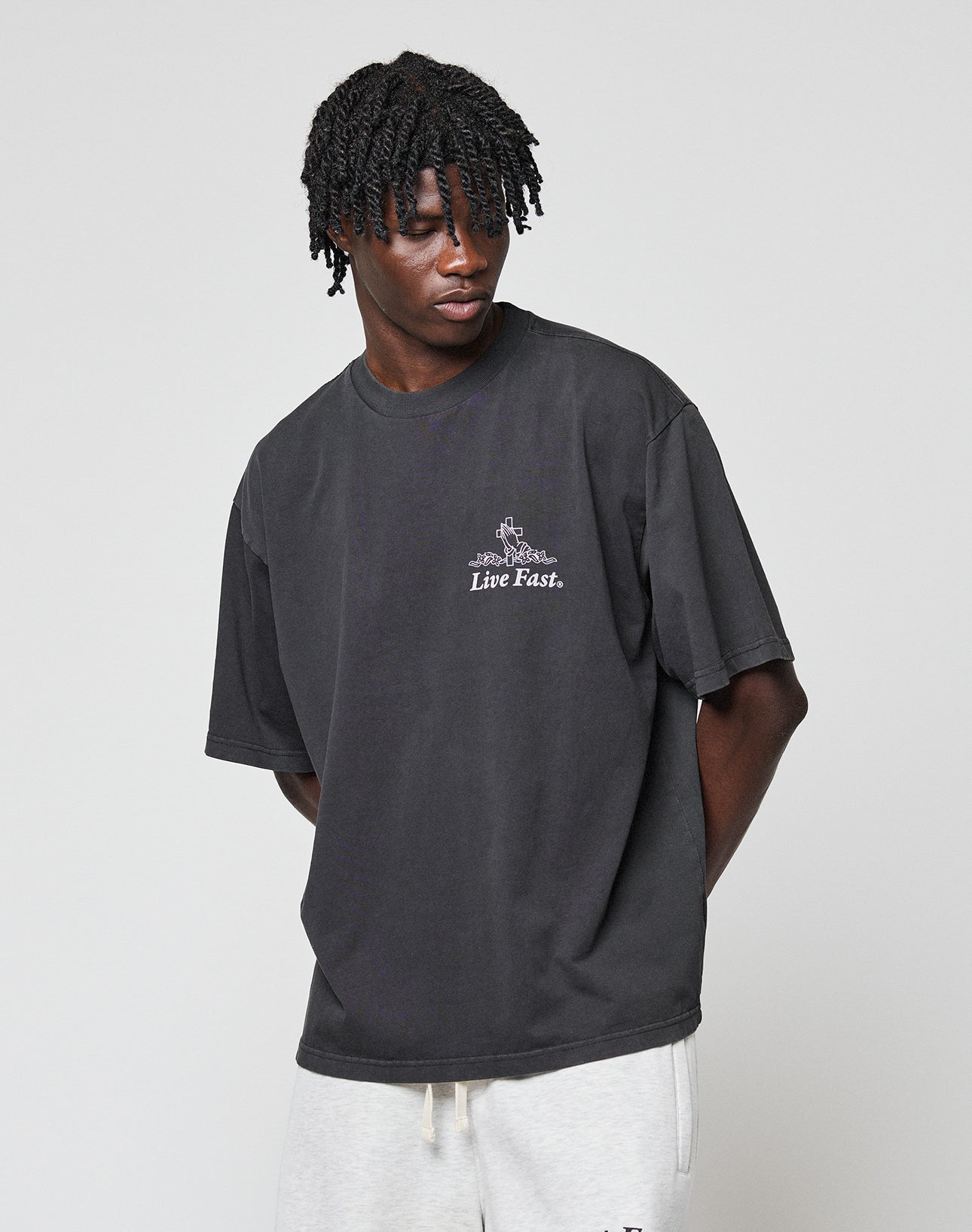 A person with short, twisted hair wears the LIV­­E FAST DIE YOUNG LFDY Praying Tee in an oversize fit with front print, paired with light gray pants, standing against a plain light background.
