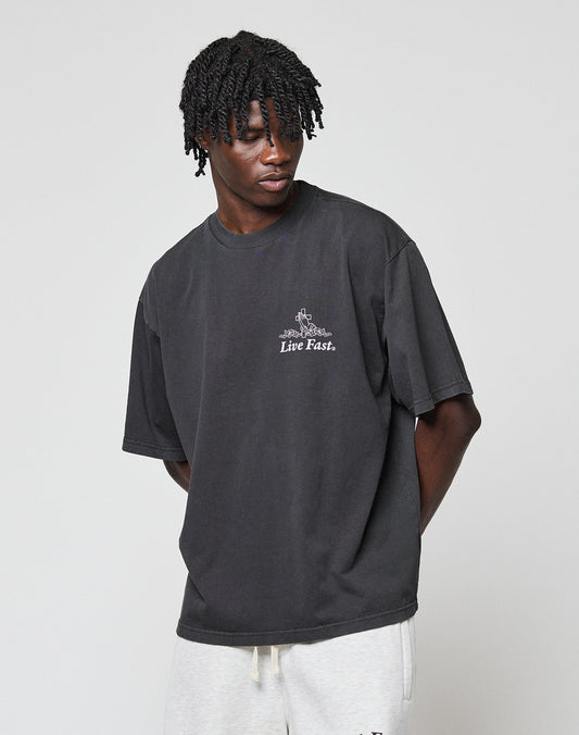 A person with short, twisted hair wears the LIV­­E FAST DIE YOUNG LFDY Praying Tee in an oversize fit with front print, paired with light gray pants, standing against a plain light background.