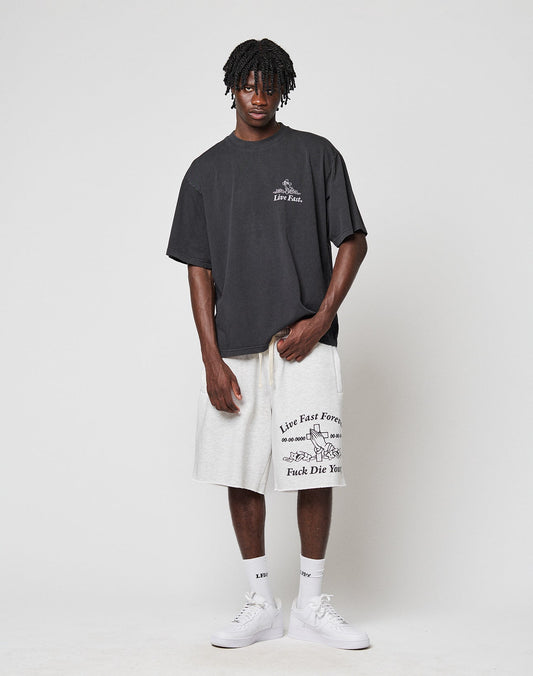 A man with short dreadlocks stands against a plain light background, wearing a loose black t-shirt and light gray LIVE FAST DIE YOUNG LFDY Praying Shorts with seitlichen Eingrifftaschen, paired with white socks and sneakers.