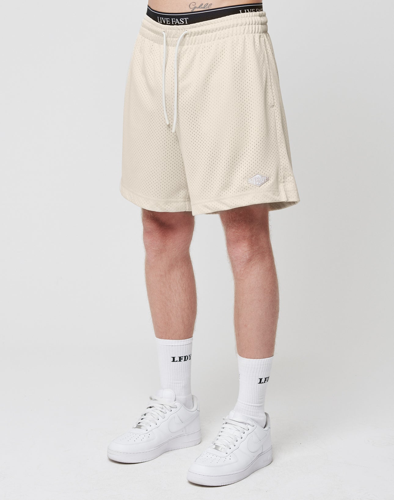 A person wears LIVE FAST DIE YOUNG Basic Mesh Shorts, LFDY-branded white socks, and white Nike sneakers, standing against a plain white background. Only the lower body is shown. | model